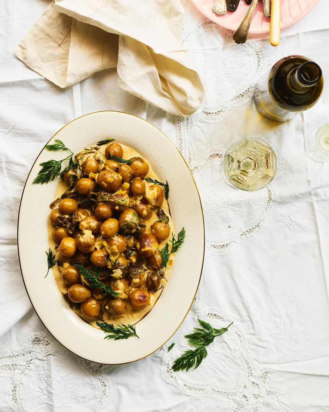 Overhead shot of creamed potatoes with herbs on a white tablecloth, alongside a wine glass and bottle, and silverware.