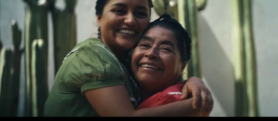 Two Hispanic women embrace indoors with smiling faces. Close-up shot showcases green and red tops, indoor setting, and happy expressions.