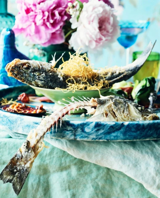 Overhead shot of cooked and partially eaten fish on decorative plates with flower arrangements in the background against a light blue wall.