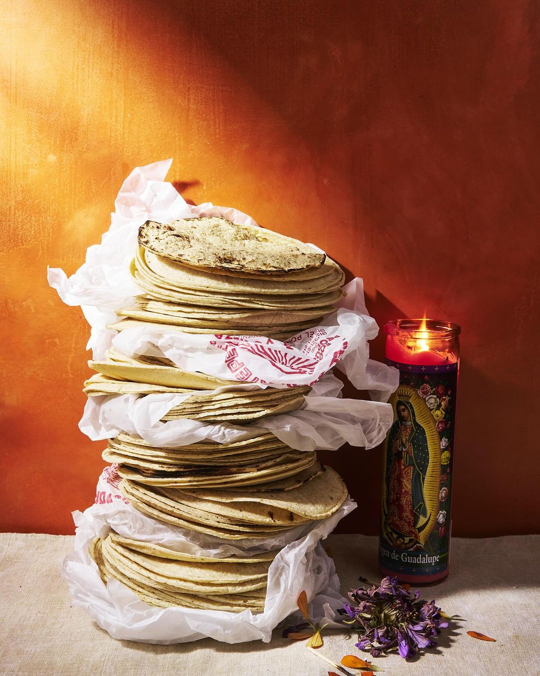 Stack of fresh corn tortillas on white tissue paper next to a religious candle against an orange textured background indoors.