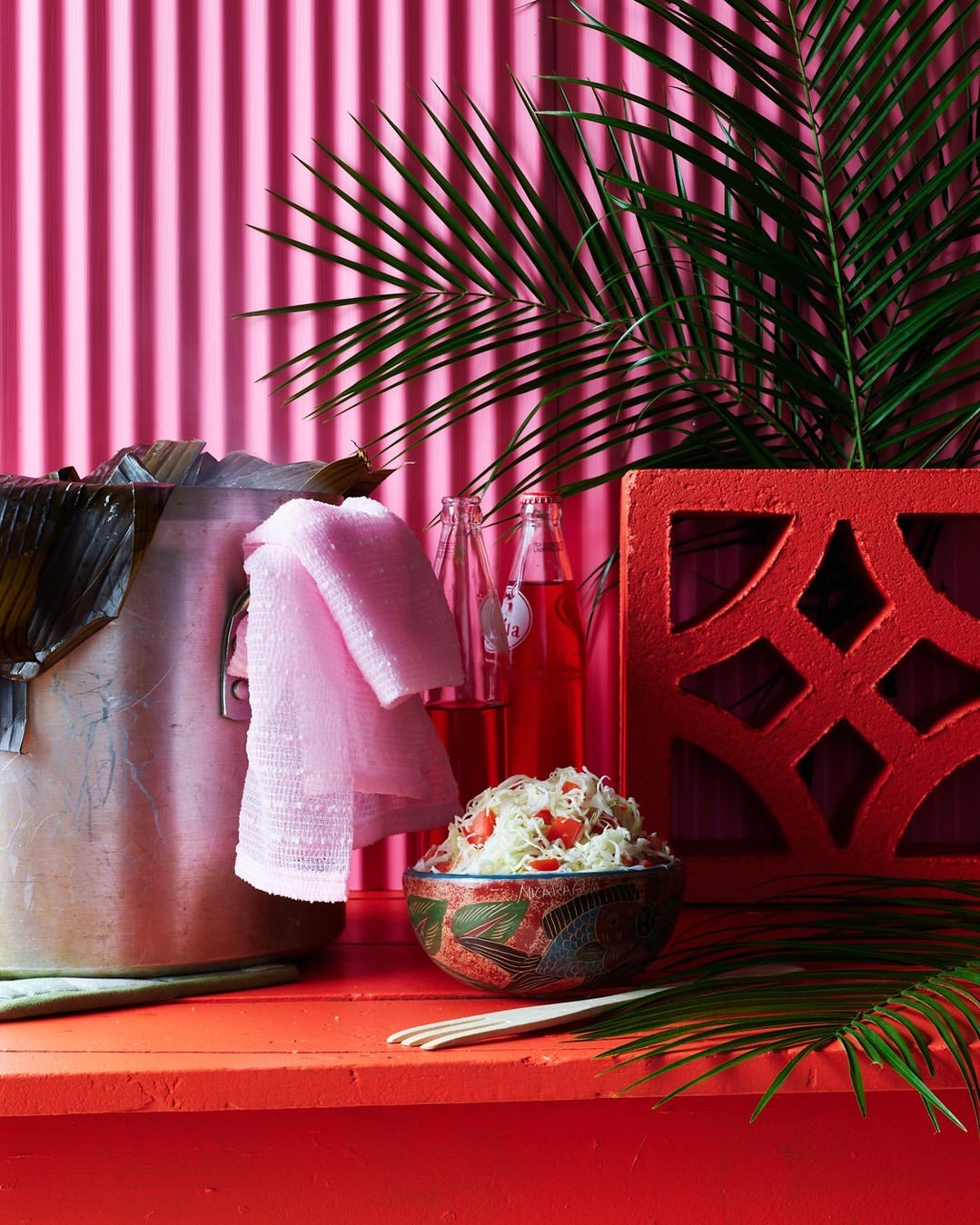 Still life photograph of a bowl of food, bottles of red liquid, and a pink towel against a pink background with palm fronds.