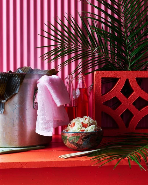 Still life photograph of a bowl of food, bottles of red liquid, and a pink towel against a pink background with palm fronds.