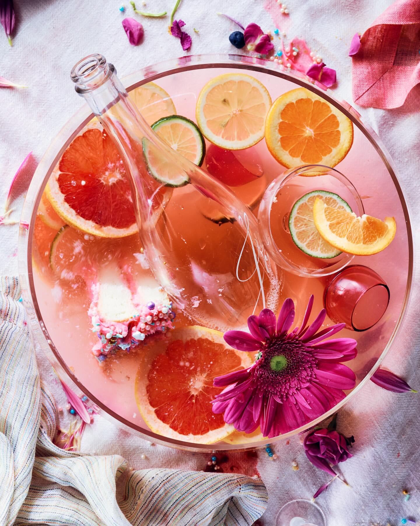 Fruit-infused beverage in a glass bowl with citrus slices, clear bottle, magenta flower, and a striped cloth background creates a vibrant, refreshing scene.