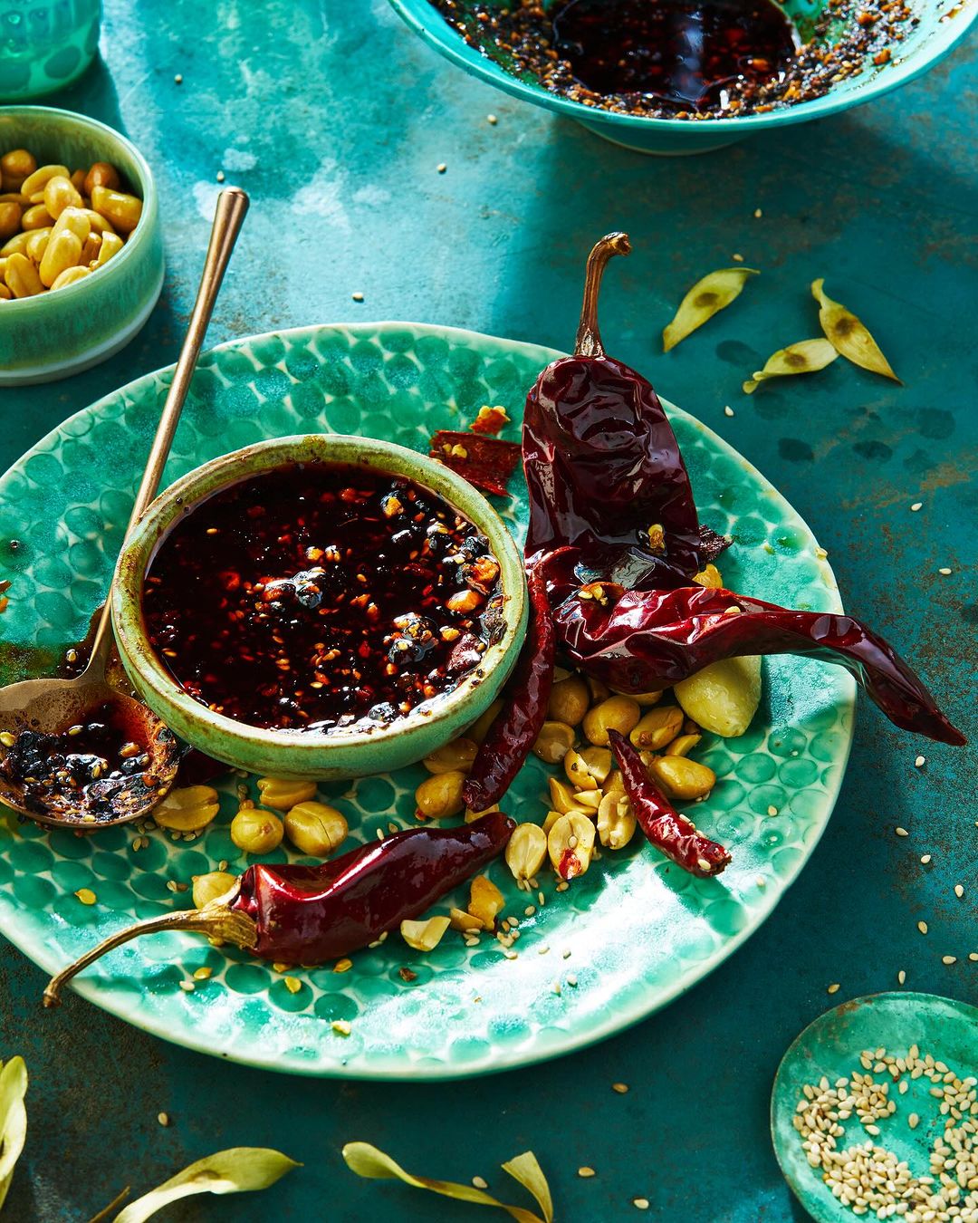 Overhead shot of a vibrant teal plate with a small bowl of dark chili oil, whole dry chilies, and peanuts, featuring a golden spoon.