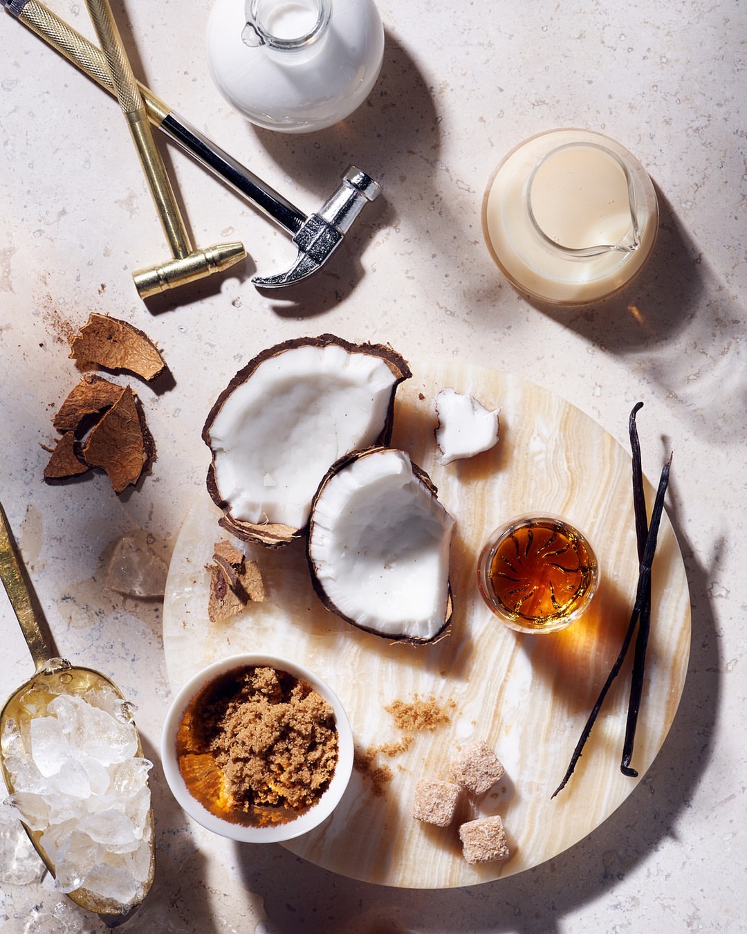 Overhead shot of halved coconut, vanilla beans, sugar cubes, ice, and bar tools on a round marble board set against a textured backdrop.