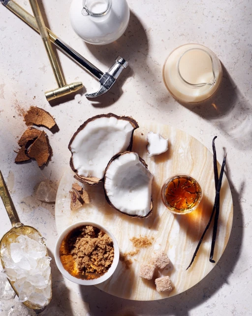 Overhead shot of halved coconut, vanilla beans, sugar cubes, ice, and bar tools on a round marble board set against a textured backdrop.