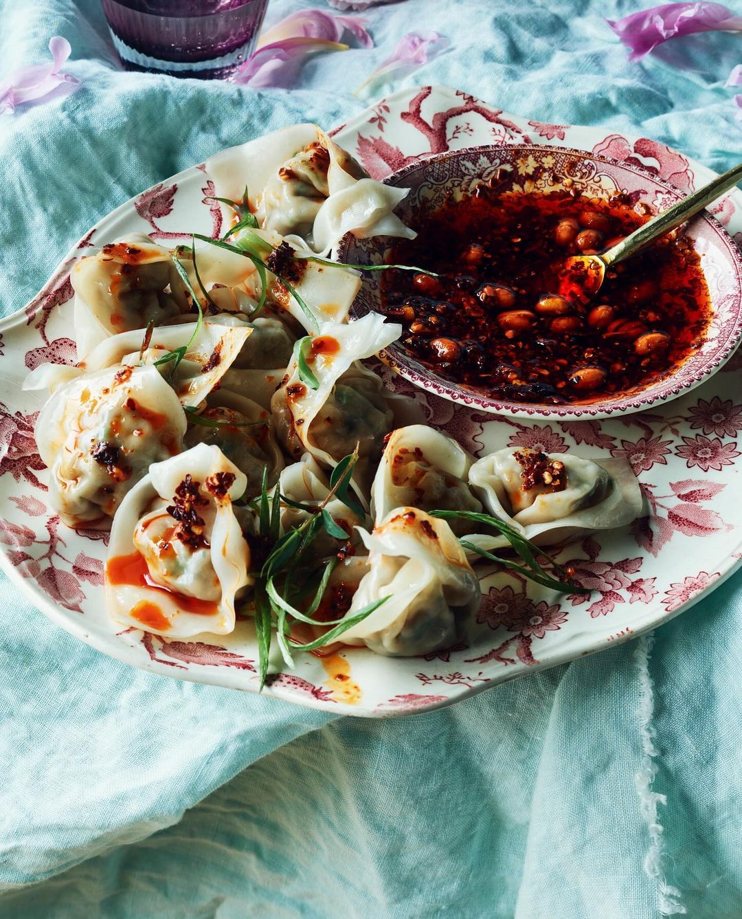 Overhead shot of dumplings with chili oil on a vintage floral plate, accented with green herbs and featuring a light blue linen backdrop.