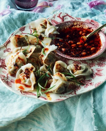 Overhead shot of dumplings with chili oil on a vintage floral plate, accented with green herbs and featuring a light blue linen backdrop.