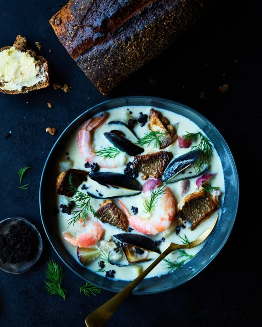 Overhead shot of seafood chowder in a blue bowl, garnished with dill, accompanied by dark bread and a spoon on a black surface.