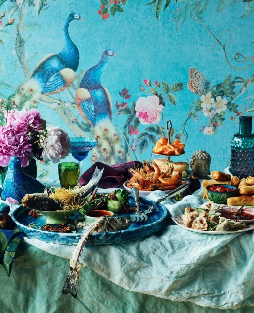 Still life arrangement showcases a variety of food items on a linen tablecloth, set against a decorative background with peacocks.