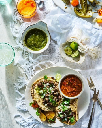 Overhead shot of tacos topped with salsa and onion, served with limes and salsa on a white fringed tablecloth.