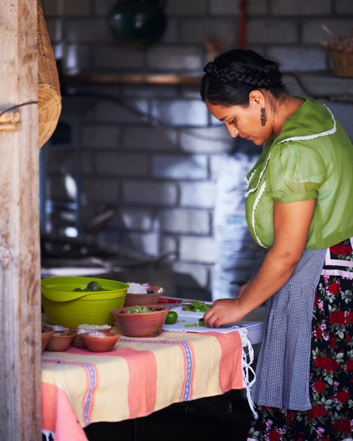 Woman in traditional blouse and floral apron prepping vegetables at a table in an outdoor kitchen with brick oven.