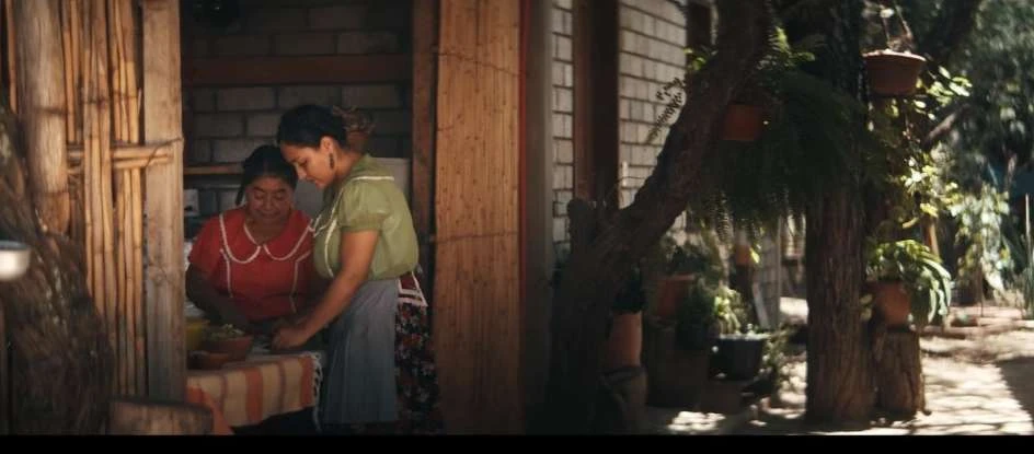 Two adult females prepare food on a table outside a rustic building with a tree in the foreground, creating a candid and cultural scene.