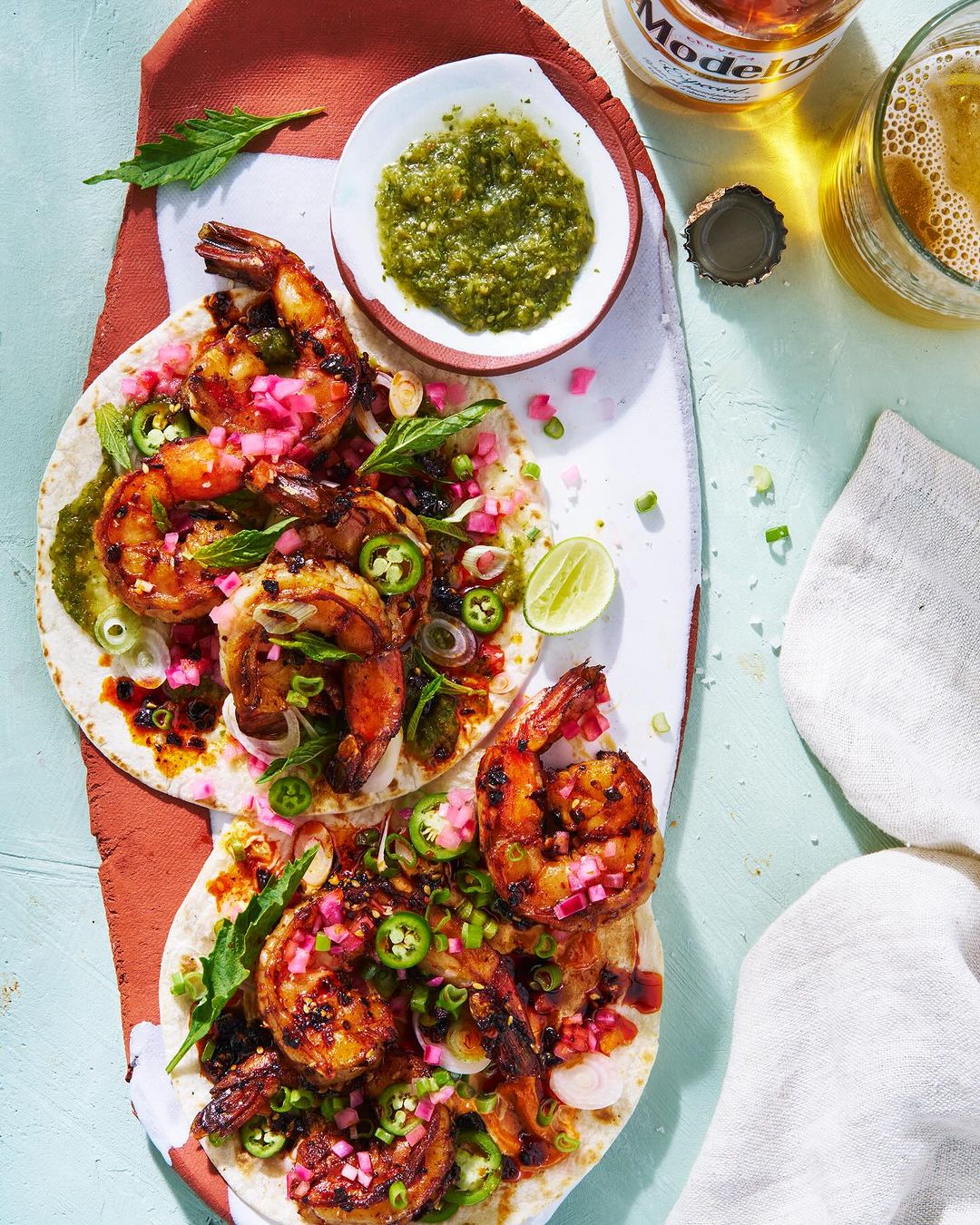 Overhead shot of grilled shrimp tacos with salsa, jalapeños, and lime wedges arranged on a white serving platter alongside a beer.