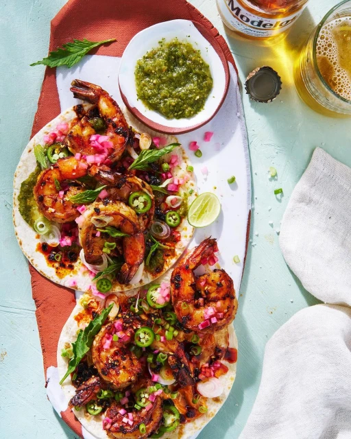 Overhead shot of grilled shrimp tacos with salsa, jalapeños, and lime wedges arranged on a white serving platter alongside a beer.