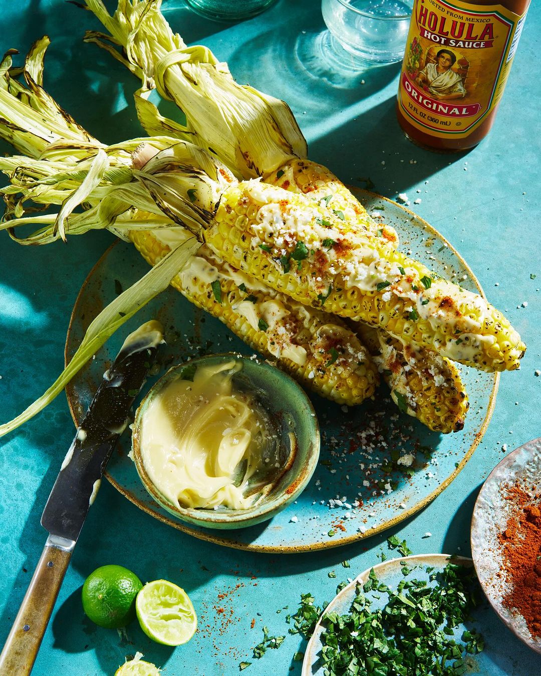 Overhead shot of grilled corn on the cob with condiments like lime, butter, spices, and Cholula hot sauce on a vibrant turquoise surface.