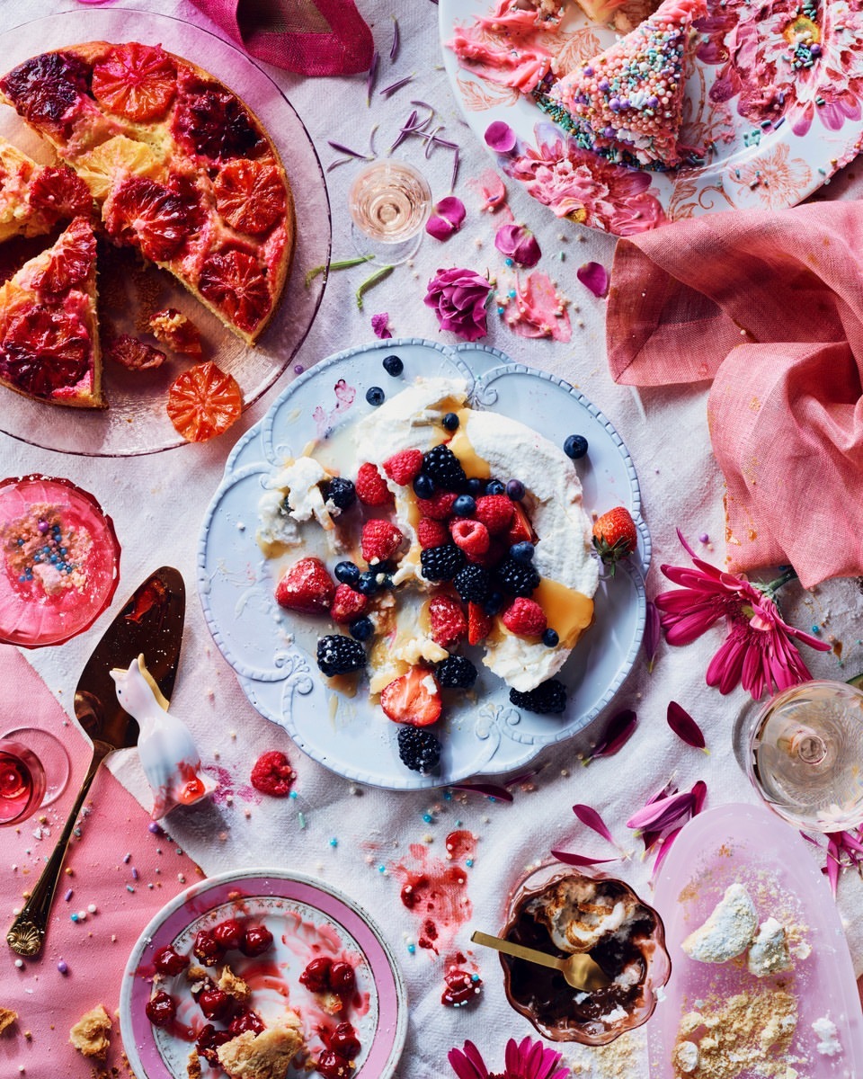 Overhead shot of dessert plates with cake, fruit, meringue, and pink flowers scattered on a light tablecloth, capturing a sweet, colorful still life.