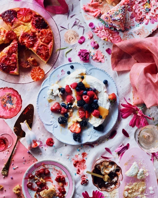 Overhead shot of dessert plates with cake, fruit, meringue, and pink flowers scattered on a light tablecloth, capturing a sweet, colorful still life.