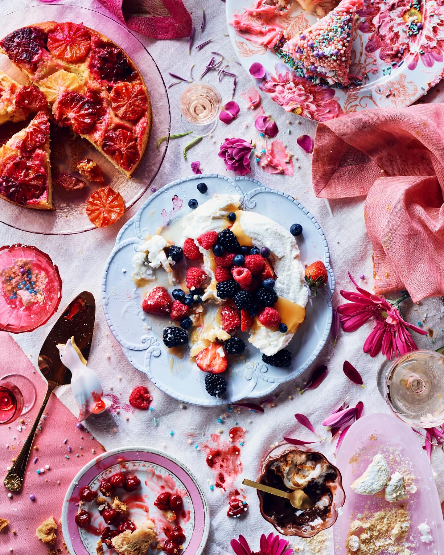 Overhead shot of assorted desserts including fruit tart, berry pavlova, cherry pie, and chocolate mousse on a decorated tablecloth.