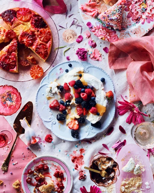 Overhead shot of assorted desserts including fruit tart, berry pavlova, cherry pie, and chocolate mousse on a decorated tablecloth.