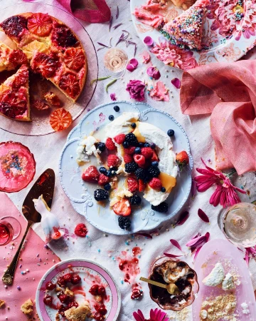 Overhead shot of assorted desserts including fruit tart, berry pavlova, cherry pie, and chocolate mousse on a decorated tablecloth.