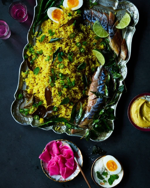 Overhead shot of plated rice dish with fish, egg, and lime on a decorative silver platter, with colorful garnishes and dark background.