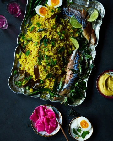Overhead shot of plated rice dish with fish, egg, and lime on a decorative silver platter, with colorful garnishes and dark background.