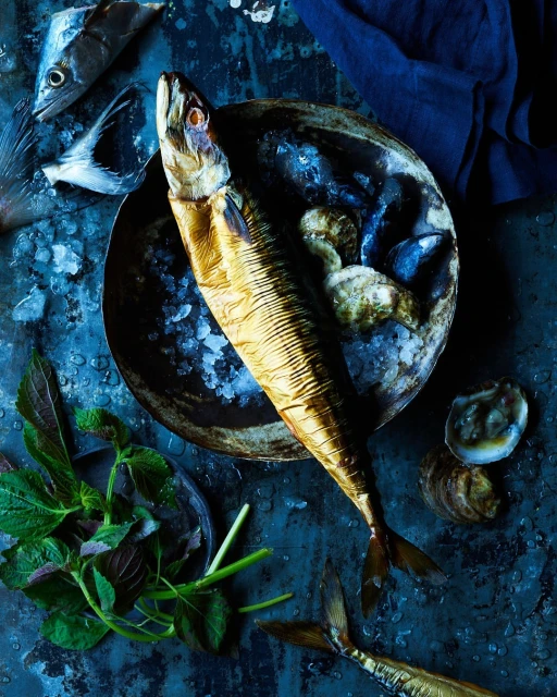 Overhead shot of arranged smoked fish, oysters, and fresh herbs on a rustic blue surface, showcasing culinary ingredients.