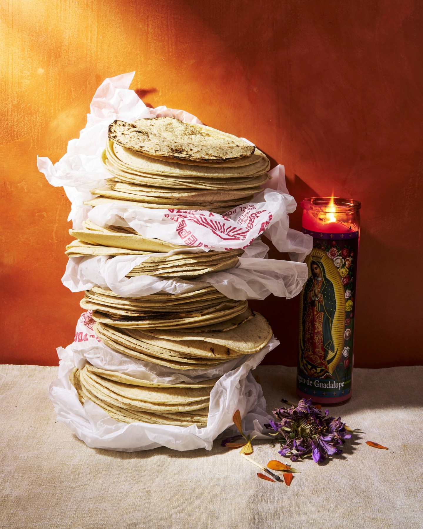 A stacked arrangement of tortillas in paper, accented by a candle and dried flowers, set against an orange backdrop.