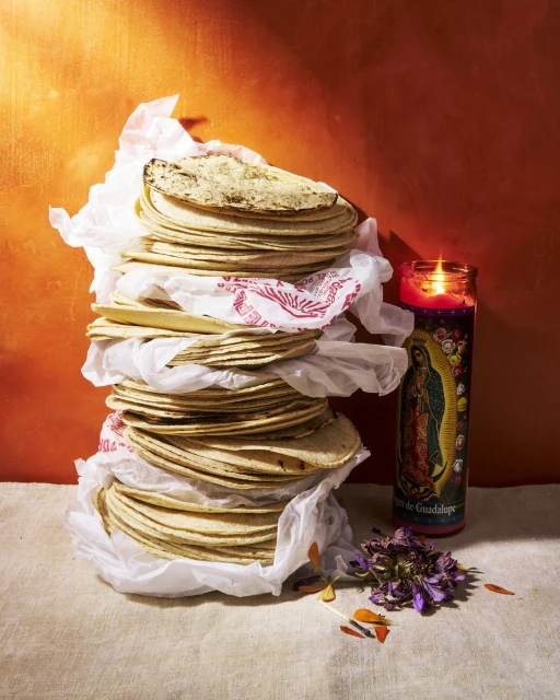 A stacked arrangement of tortillas in paper, accented by a candle and dried flowers, set against an orange backdrop.