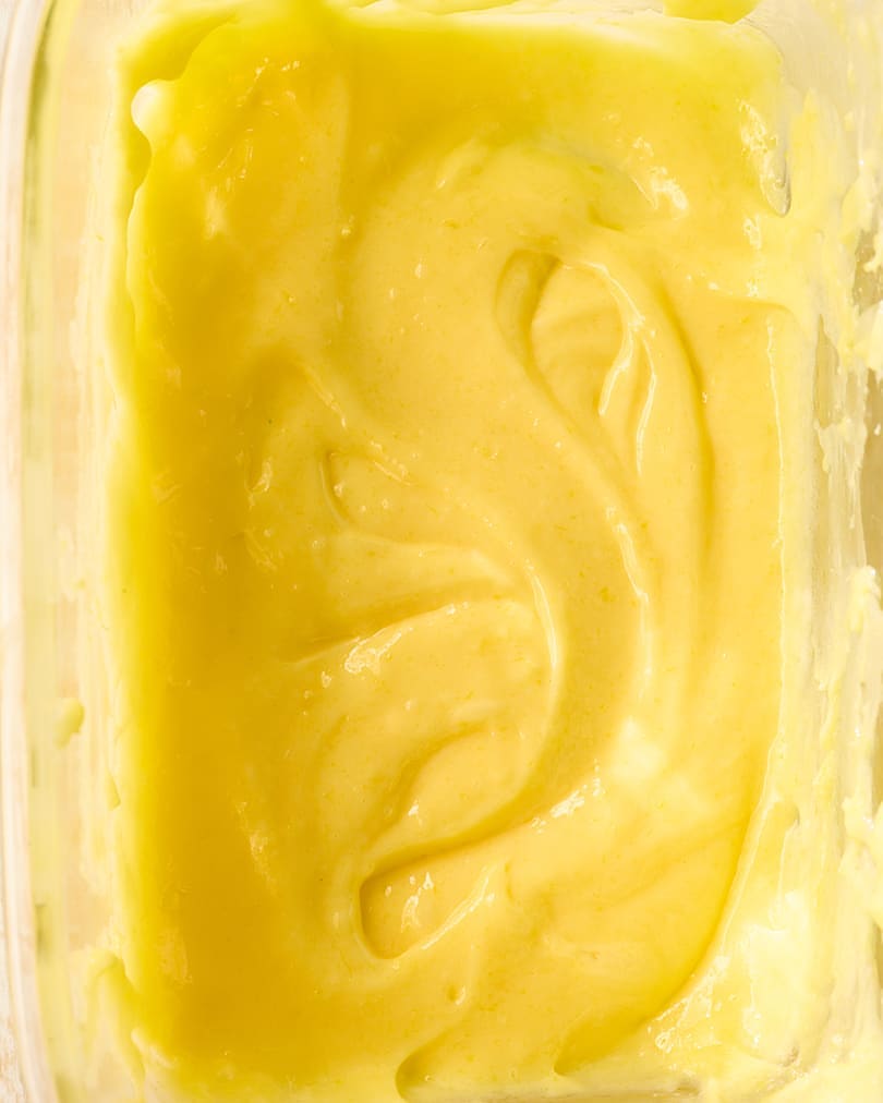 Close-up of a smooth, creamy, light yellow substance, possibly a sauce or dessert, swirled in a clear container. Food photography.