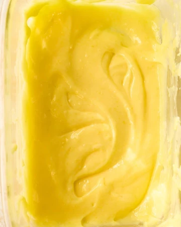 Close-up of a smooth, creamy, light yellow substance, possibly a sauce or dessert, swirled in a clear container. Food photography.