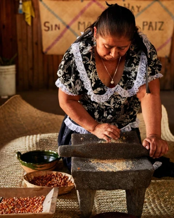 Woman grinding grain on metate with mano in traditional indoor setting. Straw mat, wooden walls, bowl of grain in the background.