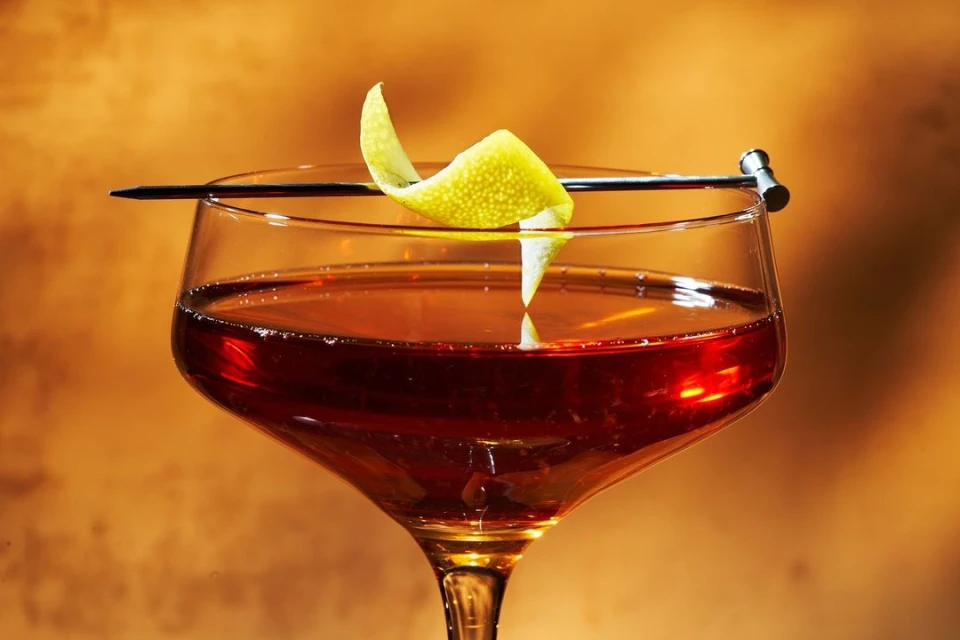 Close-up of a coupe glass filled with a dark amber cocktail, garnished with a lemon twist on a metal pick against an amber background.