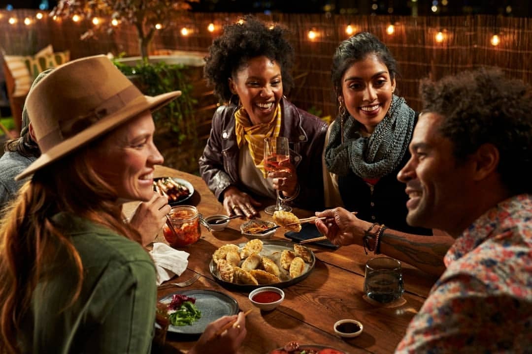 Diverse group of friends enjoys an outdoor dinner party with dumplings, salads, kimchi, and wine, illuminated by string lights at night.