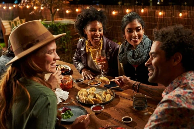 Diverse group of friends enjoys an outdoor dinner party with dumplings, salads, kimchi, and wine, illuminated by string lights at night.
