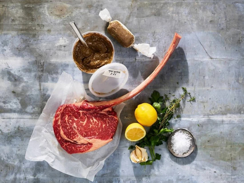 Overhead shot of a raw tomahawk steak on white parchment paper surrounded by lemon, garlic, herbs, butter, and sauce.