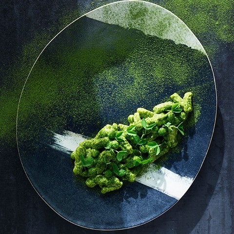 Overhead shot of green pasta dish with herbs on a navy blue and white speckled plate, sprinkled with green powder.