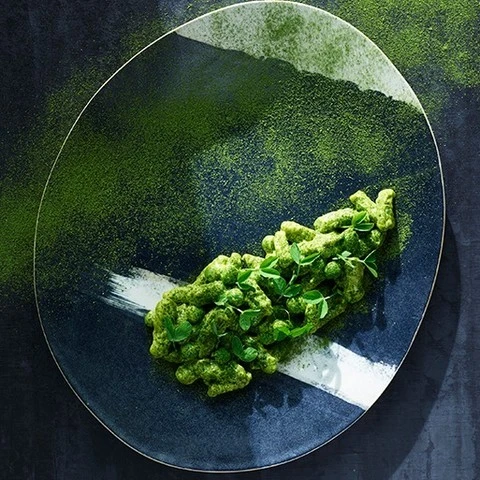 Overhead shot of green pasta dish with herbs on a navy blue and white speckled plate, sprinkled with green powder.