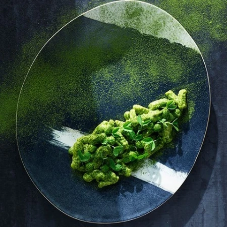 Overhead shot of green pasta dish with herbs on a navy blue and white speckled plate, sprinkled with green powder.