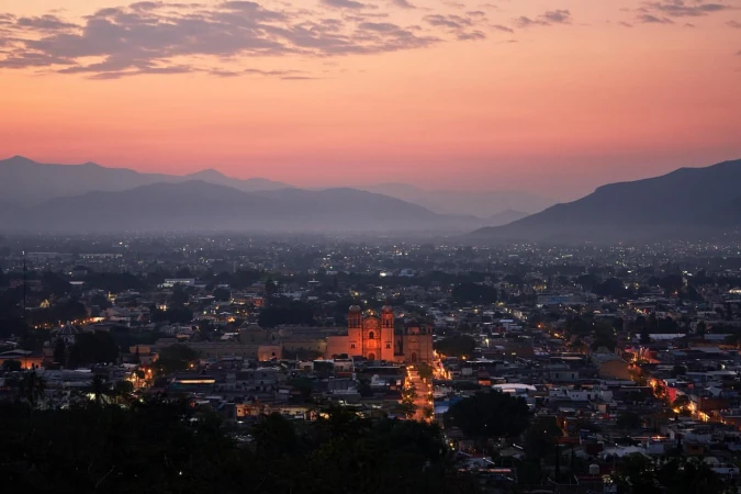 Aerial view of a city illuminated at dusk with a prominent cathedral lit up and mountain ranges silhouetted against a sunset sky.