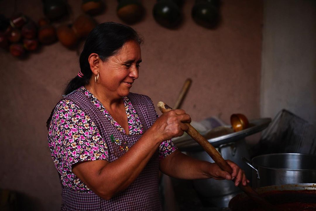 Elderly Hispanic woman stirring pot in kitchen, wearing floral shirt and apron, in traditional rural setting with hanging gourds.