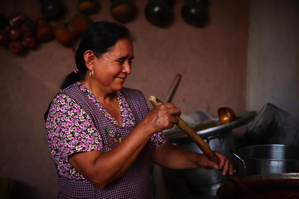 Elderly Hispanic woman stirring pot in kitchen, wearing floral shirt and apron, in traditional rural setting with hanging gourds.