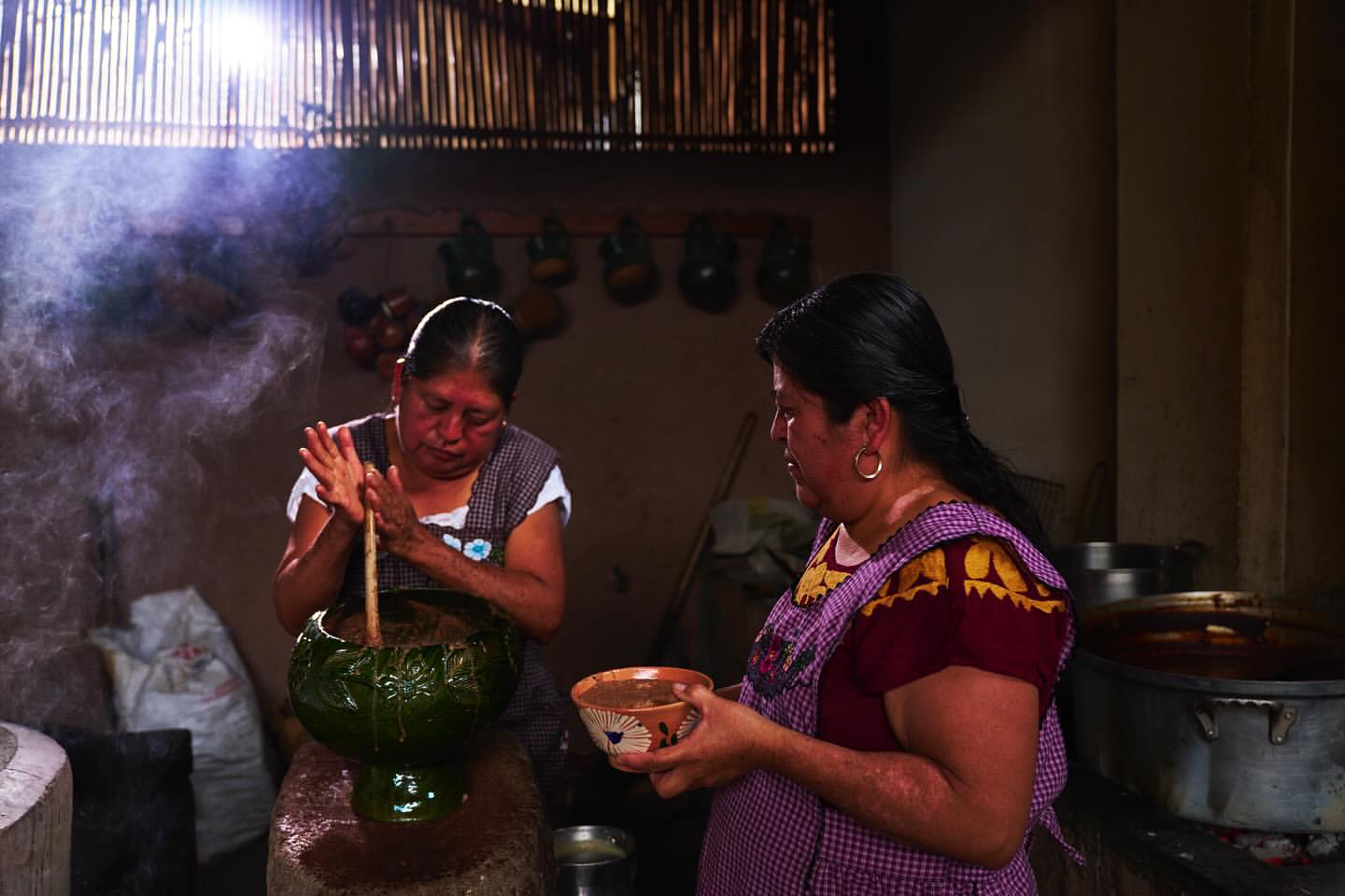 Two women in a rustic kitchen prepare a meal, one stirring ingredients in a green bowl, the other holding a bowl with a smile.