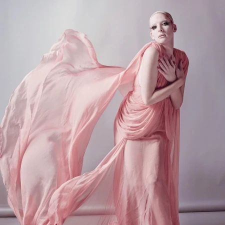 Model in a flowing pink chiffon dress with a dramatic billowing train captured against a neutral grey studio backdrop.