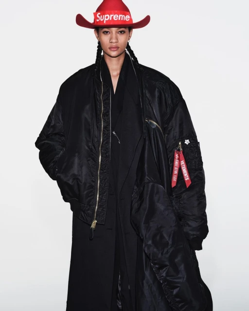Fashion shot of a model with a red cowboy hat with Supreme logo, wearing an oversized black bomber jacket in a photo studio.