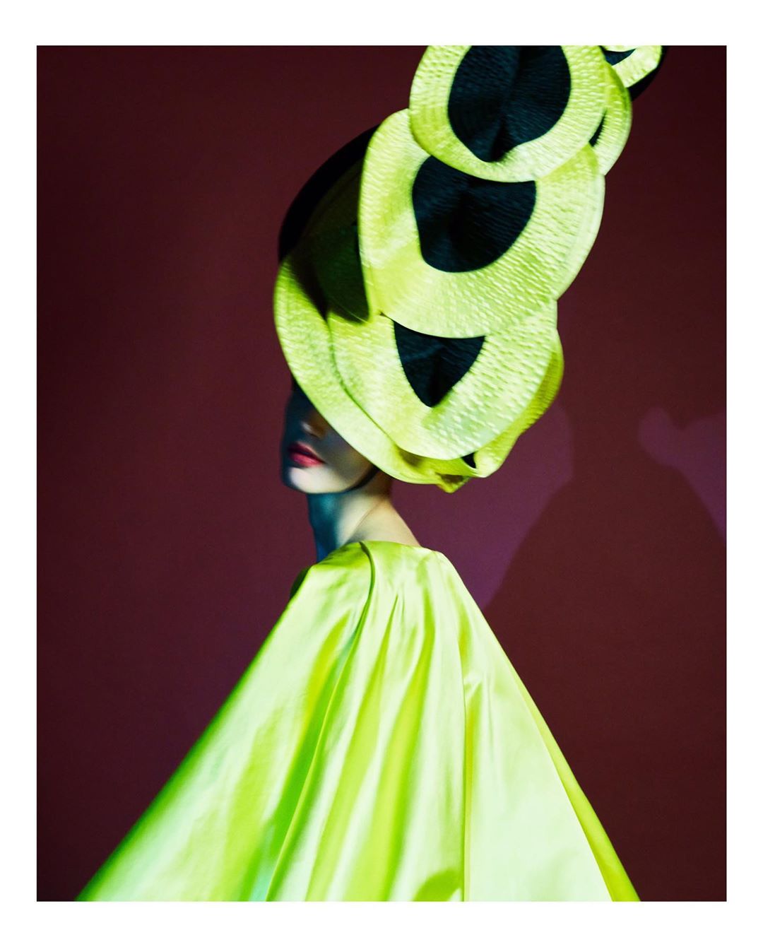 Fashion shot of a model wearing a large yellow and black hat with a matching dress against a dark red studio background.