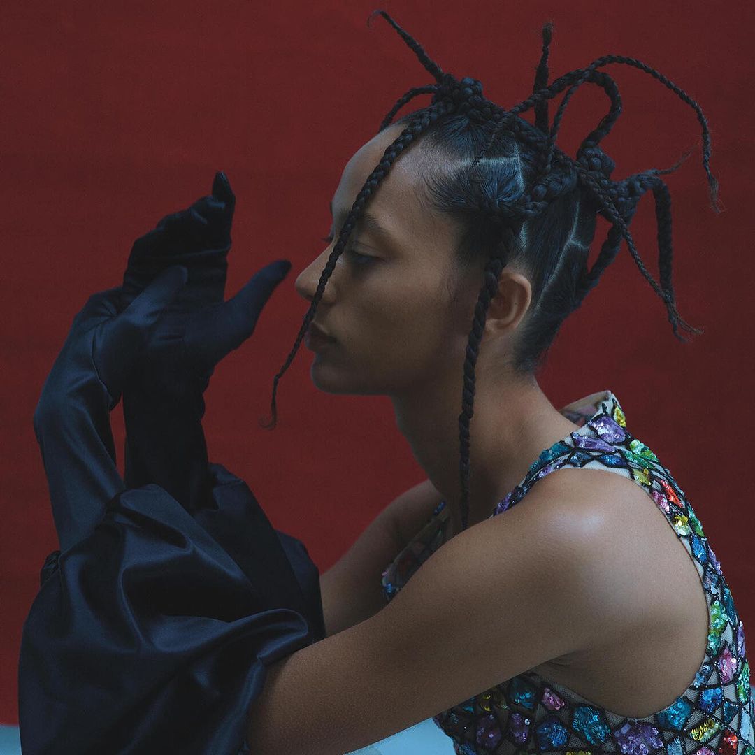 Profile view of a woman with braided hair wearing a colorful jeweled top and long satin gloves posing against a red backdrop.
