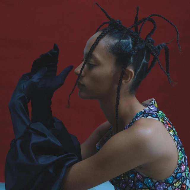 Profile view of a woman with braided hair wearing a colorful jeweled top and long satin gloves posing against a red backdrop.