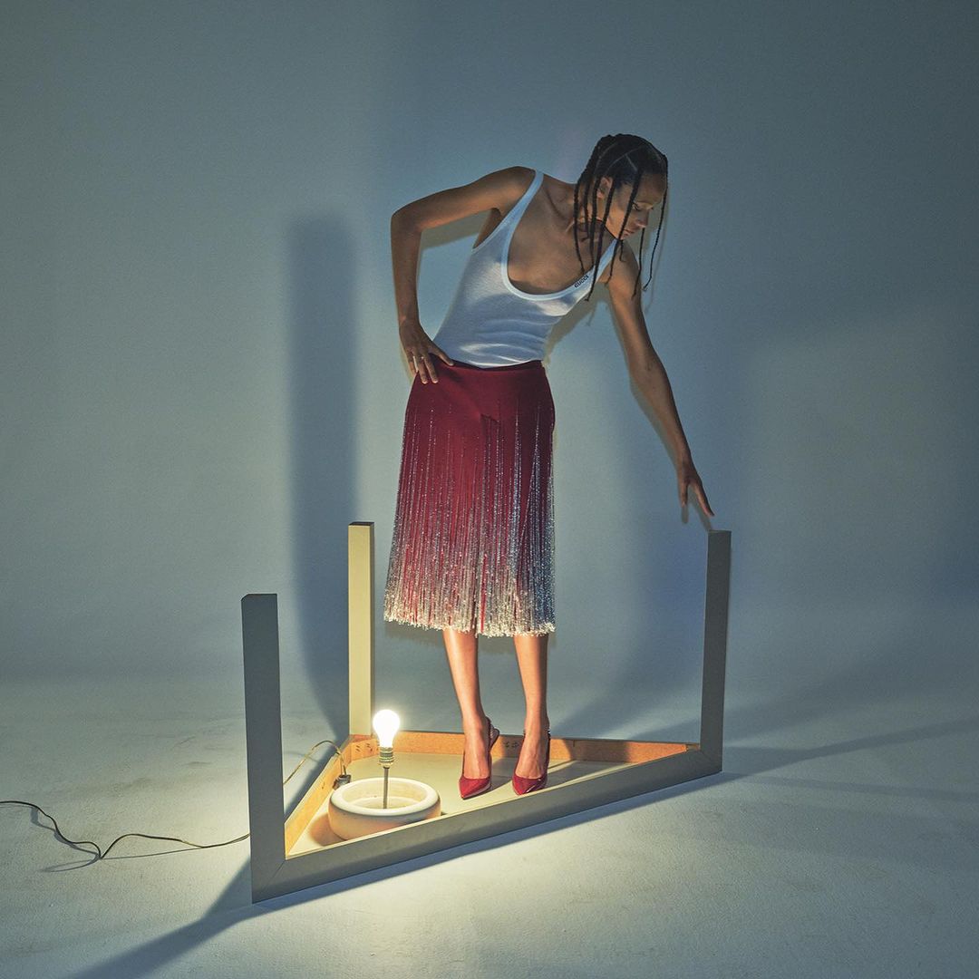 Model wearing a white tank top and red fringed skirt stands inside a triangular wooden frame with a lightbulb in a studio setting.
